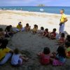 Gruppo di bambini seduti in cerchio sulla sabbia con un adulto. Spiaggia e mare sullo sfondo.