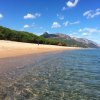 Spiaggia sabbiosa con mare calmo e montagne verdi sullo sfondo sotto un cielo azzurro.