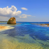 Spiaggia con mare limpido e una formazione rocciosa. Cielo azzurro con nuvole.
