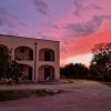 Edificio con arcate al tramonto, cielo rosa e viola, alberi e strada sterrata.